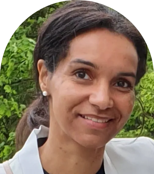 Porträtfoto einer lächelnden Frau mit dunklen Haaren, nach hinten gebunden, in weißer Bluse vor grünem, natürlichem Hintergrund.